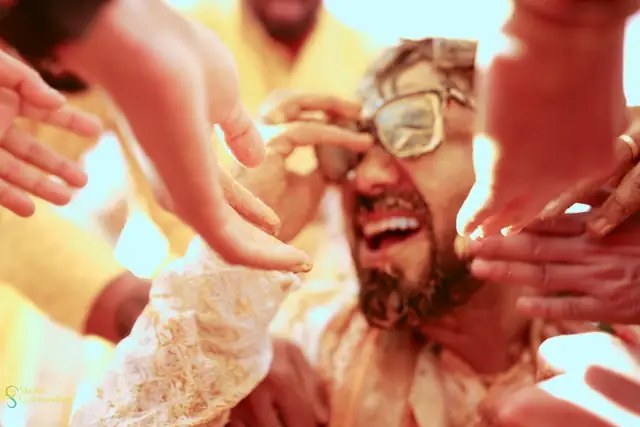 A close-up of a laughing groom in glasses, several hands reaching in to smear haldi across his face.