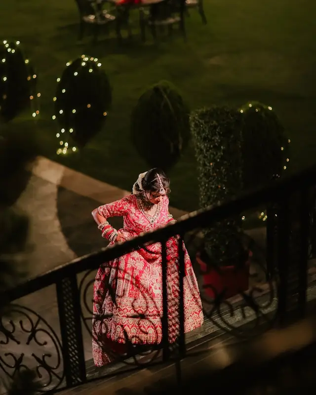 A bride in a red and pink lehenga leaning on an iron railing in a garden strung with fairy lights at dusk.