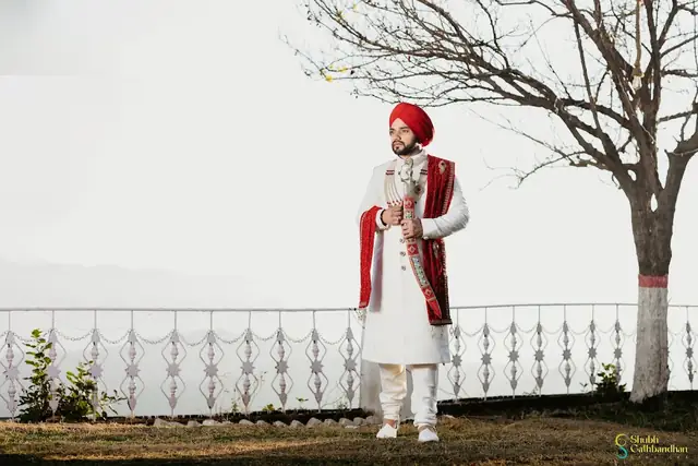 A groom in a cream sherwani with a red shawl and a red turban, standing by a railing with a bare tree and hazy hills behind him.