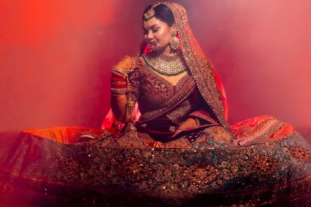 A bride in a maroon and gold lehenga seated with her skirt spread wide, red light and haze behind her.