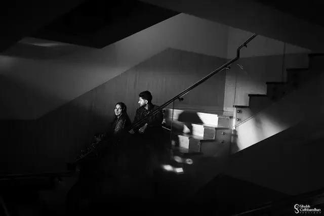 A black and white frame of a couple seated together on a stairwell landing, hard light falling across the steps.