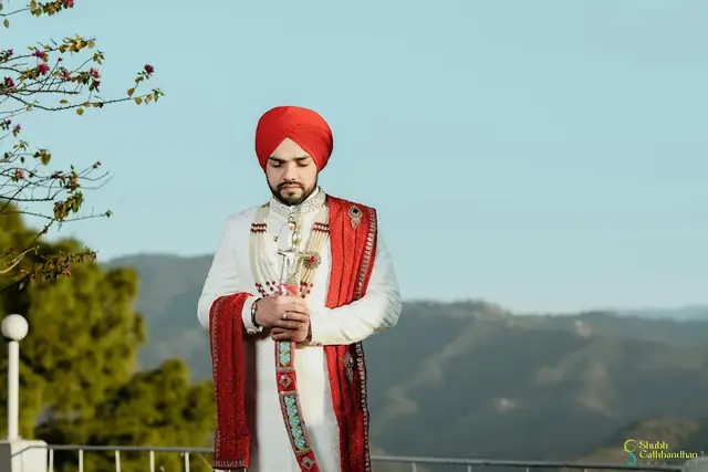 A groom in a red turban and cream sherwani with a red shawl, a wide green valley and sky behind him.